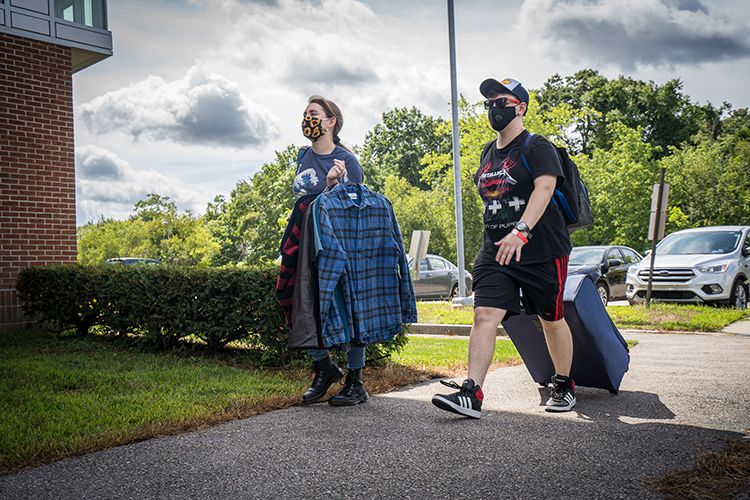 Wearing masks, two students carry clothes and a suitcase next to a residence hall.
