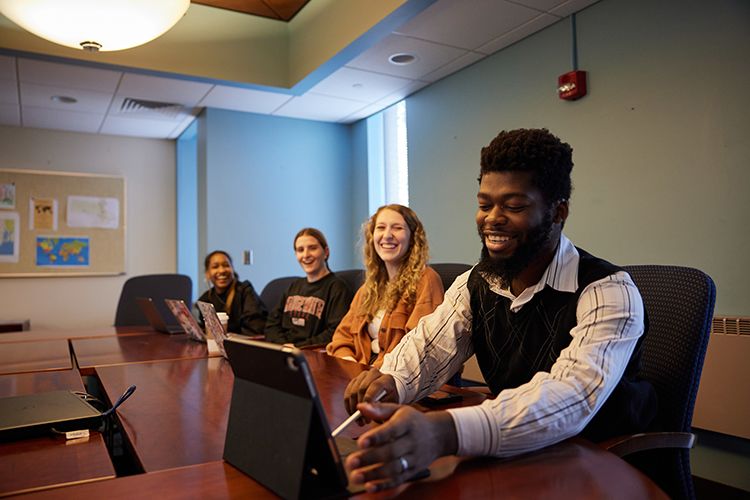 Four students talk while sitting on one side of a conference table. One of the students uses a tablet. 