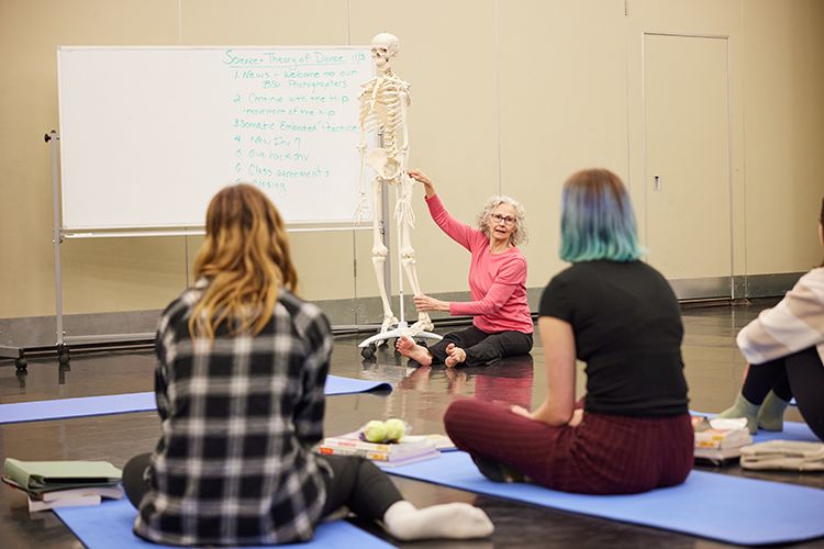 Sitting on the floor, a professor points to the leg of a skeleton. Three students sitting in the foreground watch the professor.