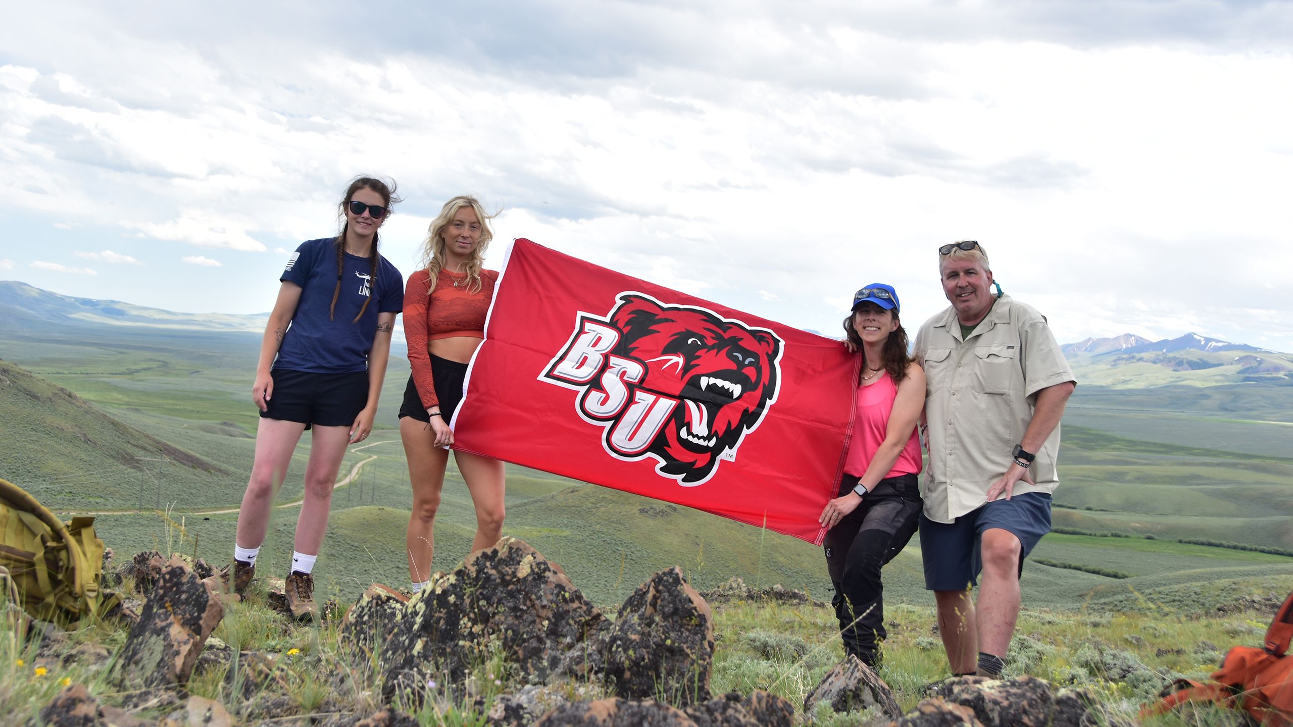 Four people hold a red BSU flag with a bear logo on top of a hill with mountains in the background.