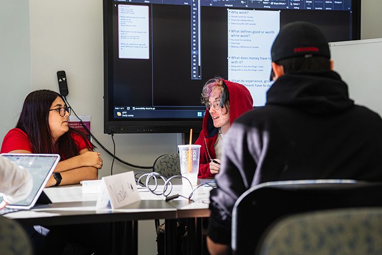 In a high-tech classroom, three students work together at a table with a large monitor on the wall behind them.