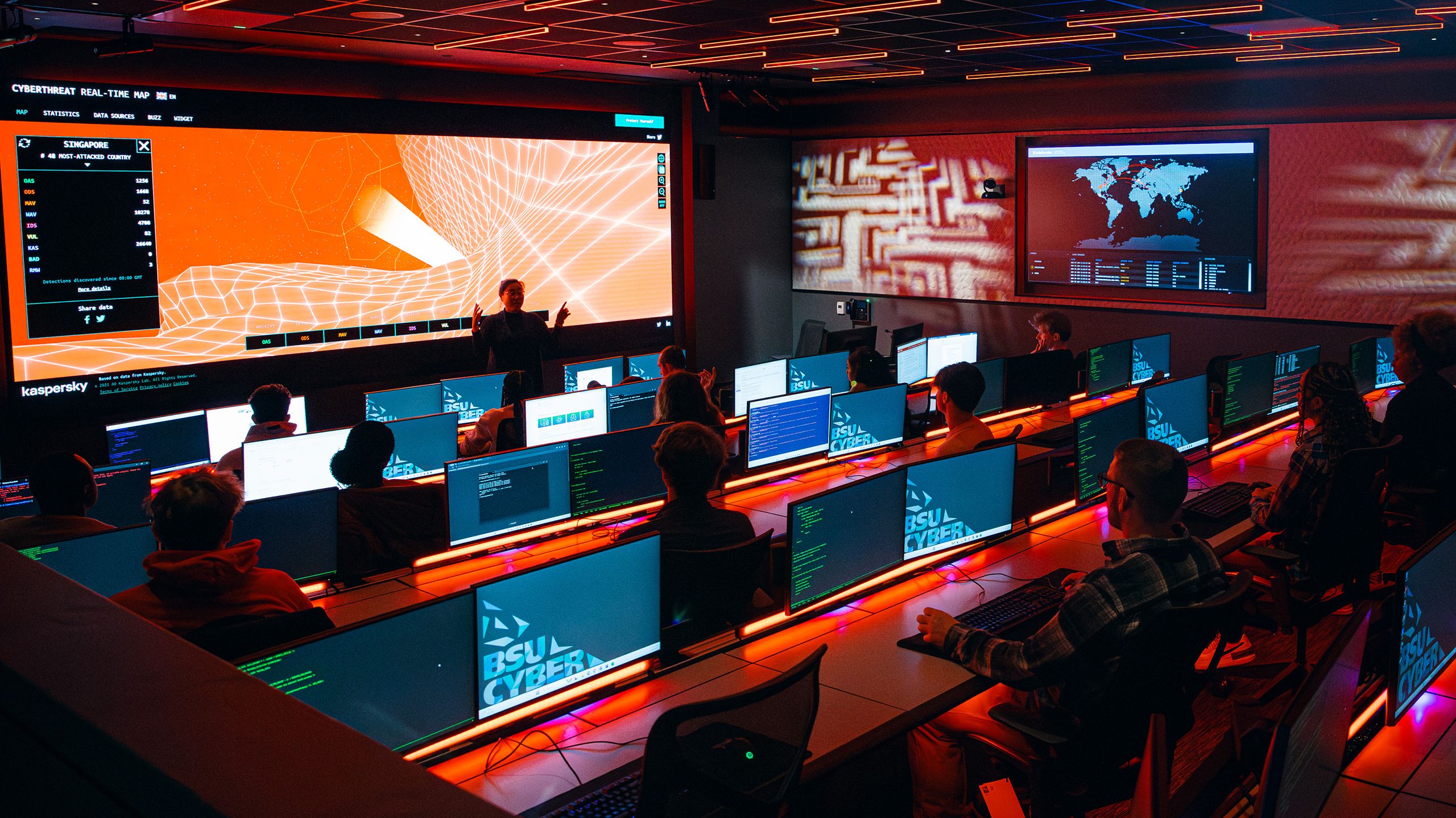 In the cyber range, students sitting behind computers listen to a professor teaching from the front of the room.