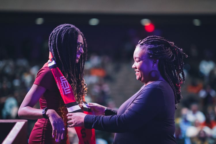 On an auditorium stage, a staff member drapes a stole around a student’s neck.
