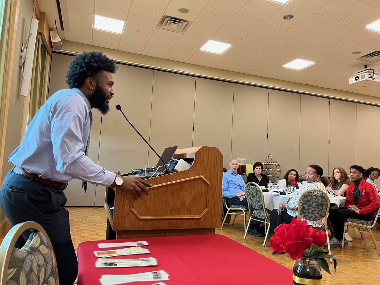 Standing behind a podium, Joseph Clark speaks to people sitting at round tables.