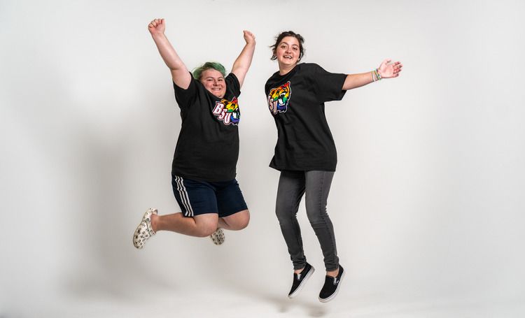 Two students wearing BSU rainbow bear t-shirts jump in front of a white background.