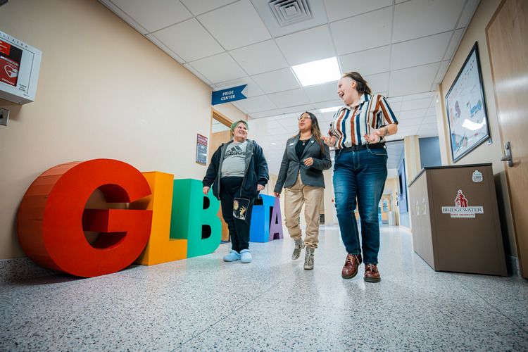 Three people walk down a hallway and pass large rainbow-colored GLBTA letters.