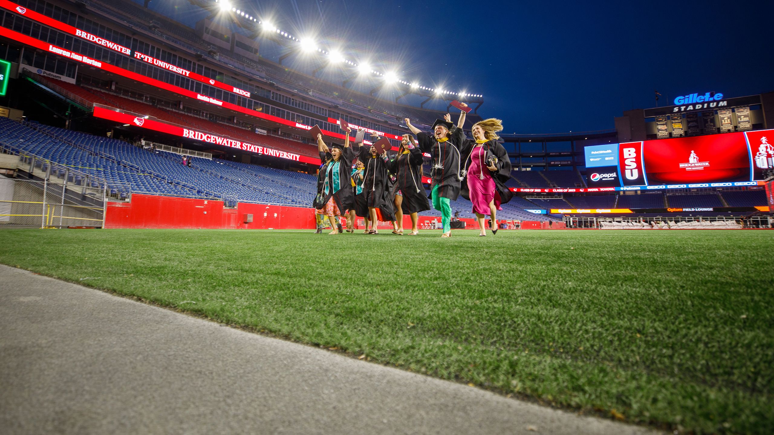 Wearing caps and gowns, several graduates run excitedly across the Gillette Stadium field.