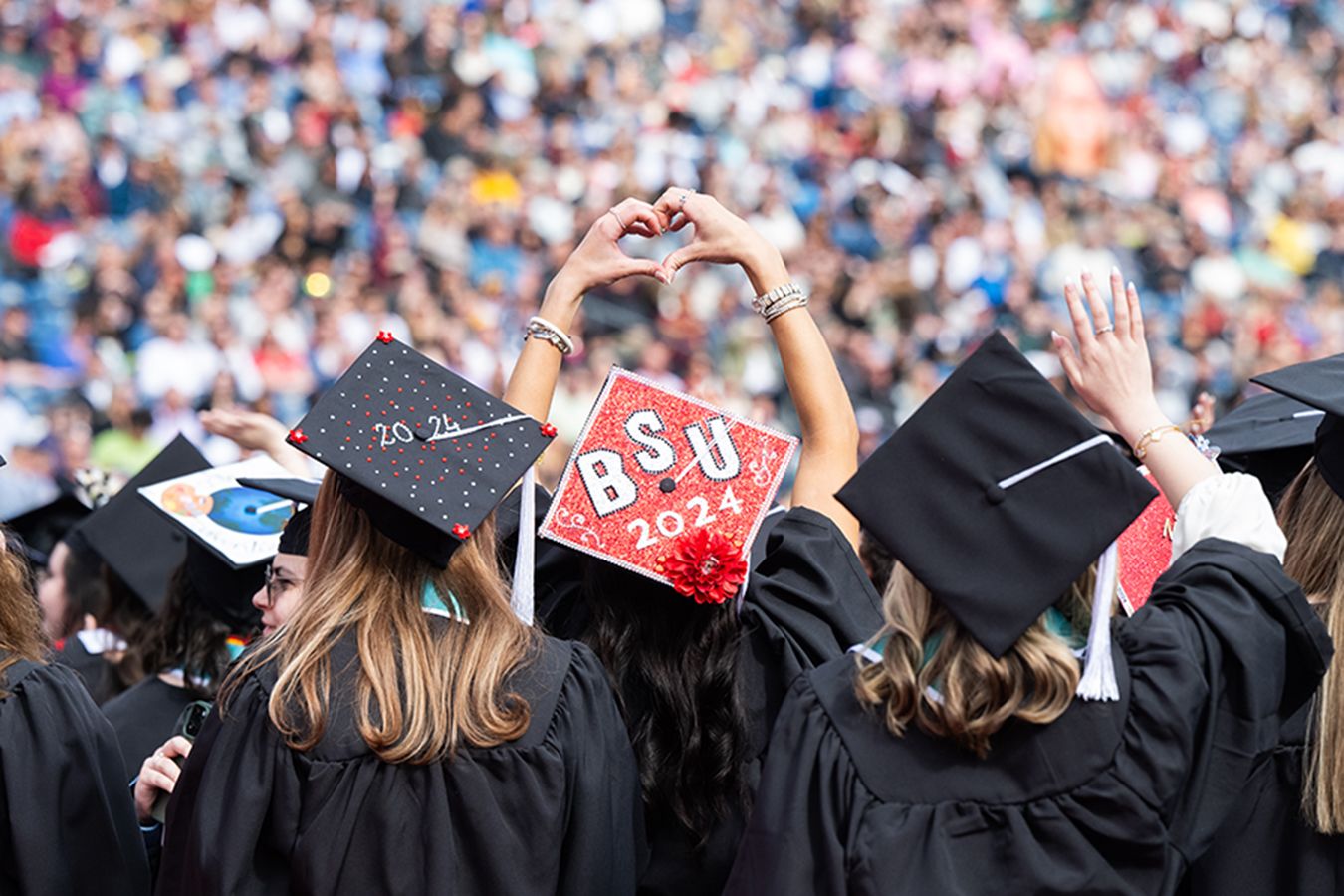 During commencement, a student wearing a BSU 2024 cap faces the audience and makes a heart sign.